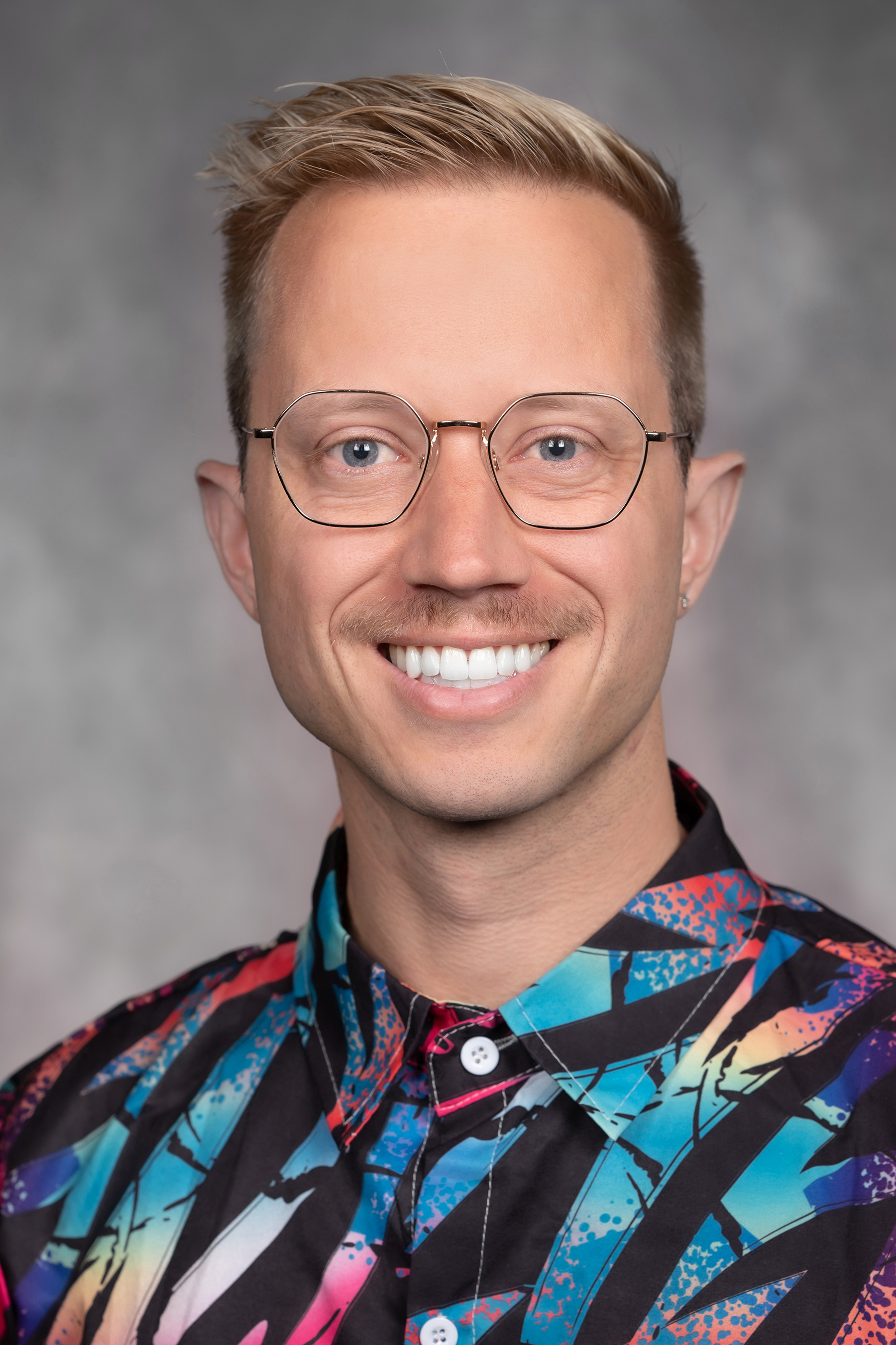 Portrait of Brady Johnson, MD MPHTM, smiling in gold-rimmed glasses and a colorful patterned shirt.